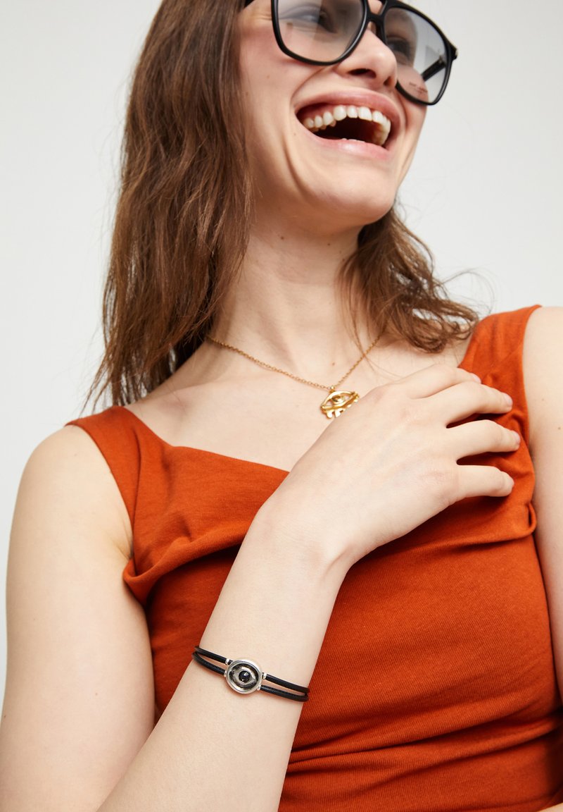 Pulsera de cuero negro con herrajes circulares de plata y un pequeño detalle negro, llevada en una mano contra una blusa de tela naranja.