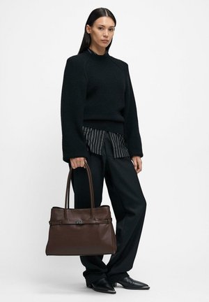 Donna che indossa un maglione lavorato a maglia nero, camicia a righe, pantaloni neri larghi e scarpe nere appuntite che tiene in mano una grande borsa tote in pelle marrone.