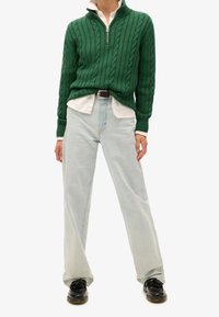 Vihreä palmikkoneule, jossa on vetoketjullinen kaulus, valkoisen paidan päällä. Vaaleansiniset leveälahkeiset farkut ja mustat kengät täydentävät asun.