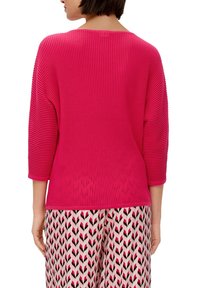 Fuchsia Rippstrickpullover mit Dreiviertelärmeln, weitem Ausschnitt und lockerer Passform, kombiniert mit gemusterten rosa und schwarzen Hosen.