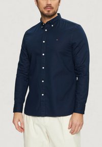 Camicia blu navy a maniche lunghe con colletto, dotata di bottoni bianchi e un piccolo logo decorativo. Realizzata in tessuto morbido, con un design aderente.