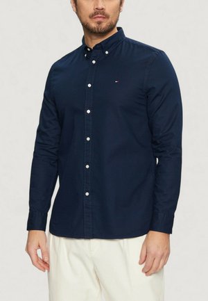 Camicia blu navy a maniche lunghe con colletto, dotata di bottoni bianchi e un piccolo logo decorativo. Realizzata in tessuto morbido, con un design aderente.