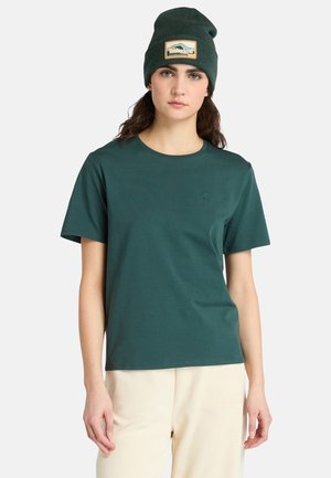Grøn bomulds t-shirt med rund hals og korte ærmer, der har et lille broderet logo på brystet. Sammen med beige bukser.