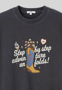 Sweatshirt gris foncé avec un design graphique incluant une botte de cowboy, des fleurs colorées et un texte ludique en blanc et orange.