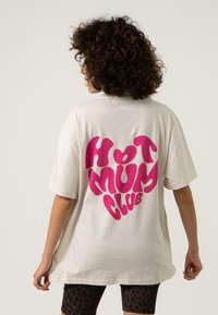 Pilkai oversized marškiniai, kuriuose ant nugaros dideliu, ryškiu rožiniu "HOT MUM CLUB" grafiniu ženklu, pagaminti iš švelnios medvilnės.