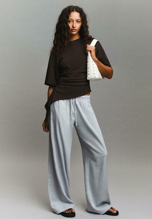 Frau mit langen lockigen Haaren, die ein schwarzes asymmetrisches Top, hellgraue weite Hosen, schwarze Sandalen trägt und eine weiße, mit Nieten besetzte Handtasche hält.