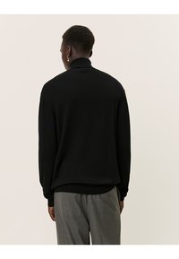 Schwarzer gerippter Rollkragenpullover mit langen Ärmeln, der eine glatte Textur und eine enganliegende Silhouette aufweist, getragen über grauen karierten Hosen.