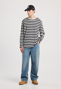 Gestreepte langemouwen blouse in marineblauw en crème, ontspannen pasvorm, gecombineerd met vervaagde blauwe wijde jeans en tan instapschoenen. Zwarte cap getoond.