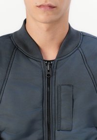 Giacca bomber blu navy con chiusura a zip, colletto a coste e una piccola tasca a toppa. Il materiale sembra liscio e leggermente lucido.