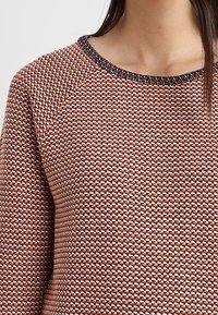 Sweter dziergany z teksturowanym wzorem diamentowym w kolorze koralowym i białym. Posiada ciemny rant przy dekolcie oraz rękawy raglanowe.