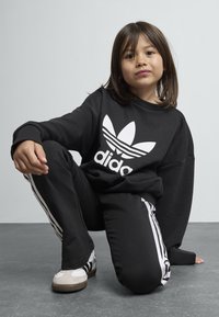 Sweatshirt preto com um corte oversized e logotipo branco, combinado com calças pretas com riscas brancas nas laterais, e ténis brancos com solas escuras.