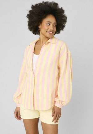 Femme aux cheveux bouclés et volumineux portant une chemise à rayures jaunes et roses boutonnée sur un haut blanc et un short jaune pâle, souriante et regardant sur le côté.