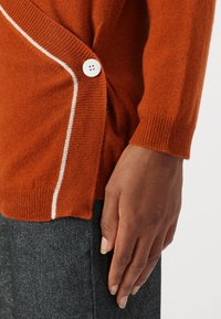 Gestrickter oranger Pullover mit einem weißen Knopfdetail und kontrastierendem Nähen entlang der Seite. Weiche Textur, taillierte Ärmel und gerippte Bündchen.