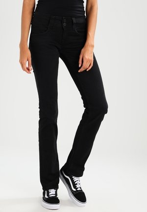 Smaltsiddende sorte jeans båret med sorte og hvide sneakers på en person, der står mod en ensfarvet lys baggrund.