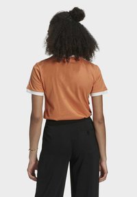 Kortärmad orange t-shirt i bomull med vit accentdetalj på ärmarna, kombinerad med svarta byxor. Baksidan visar den avslappnade passformen och materialet.