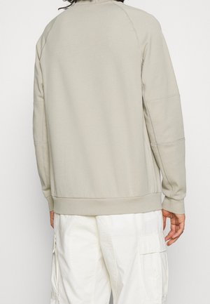 Person wearing a beige long-sleeve sweatshirt and white cargo pants, shown from the back against a plain background.