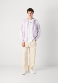Hanorac cu fermoar de culoare violet deschis, cu mansete elastice și șnururi, purtat peste un tricou alb, asortat cu pantaloni largi de culoare crem și adidași albi.