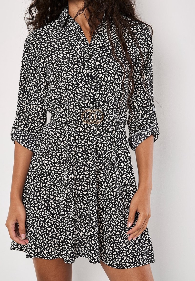 Femme portant une robe à motifs noir et blanc avec des manches retroussées et une ceinture dorée à double anneau à la taille.