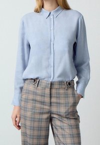 Kvinde iført en lyseblå skjorte med knapper, stoppet ind i højtaljede, beige og grå ternede bukser, med den ene hånd i lommen.