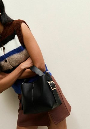 Mujer con una blusa de punto sin mangas multicolor y falda marrón, sosteniendo un bolso de cuero negro con una gran correa con hebilla.