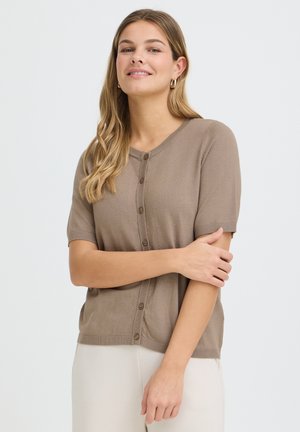 Hosszú szőke hajával rendelkező nő, aki rövid ujjú, taupe színű, gombos kardigánt és fehér nadrágot visel, karját keresztbe téve áll.