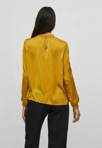 Blouse en satin jaune moutarde à manches longues, smocks texturés au col et aux poignets, avec une fermeture à boutons dans le dos. Coupe cintrée.
