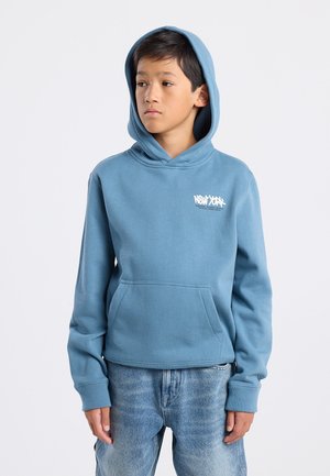 Blauwe hoodie gemaakt van fleece materiaal, met een voorkant zak en een wit tekstlogo op de borst. Gegdraagd met lichtblauwe spijkerbroek.