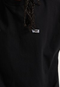 Personne portant un t-shirt Vans noir avec un logo blanc sur la poitrine et des cheveux bouclés partiellement visibles.