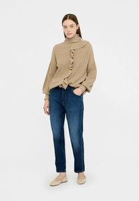 Beżowy dziany sweter z golfem i falbaną, zestawiony z niebieskimi jeansami o prostych nogawkach. Modelka nosi jasne baleriny. Proste tło.