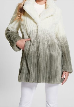 Femme portant un long manteau en fausse fourrure douce avec un dégradé allant du blanc au col au gris clair à l'ourlet, associé à un pantalon blanc.