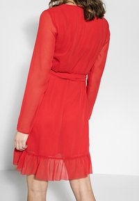 Vestido rojo transparente con mangas largas, cintura ajustada y un dobladillo acampanado con volante en la parte inferior. Textura suave y color sólido, sin estampados.