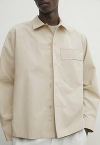 Camicia casual beige, realizzata in tessuto leggero, presenta una chiusura con bottoni, una tasca singola sul petto e una vestibilità rilassata con maniche lunghe.