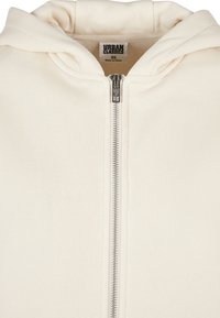 Sweat à capuche zippé blanc cassé avec fermeture éclair métallique et capuche, montrant l'étiquette de la marque Urban Classics et l'étiquette de taille XS à l'intérieur du col.