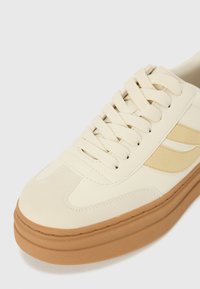 Baskets beiges avec une tige texturée, dotées de lacets crème et d'une semelle en caoutchouc beige. Présentent de subtils accents bicolores sur les côtés.
