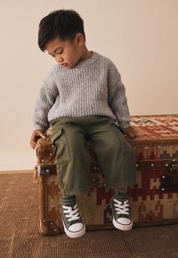 Maglione grigio a coste, pantaloni cargo verdi e sneakers alte verdi. Bambino seduto su un baule marrone decorato con superficie texturizzata.