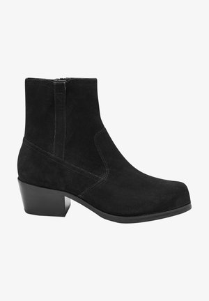 Sort ruskind ankelstøvle med en let rundet tå, blokhæle, sidelukning med lynlås og minimale syningsdetaljer på skaftet.