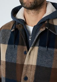 Pruun ja meriin blauhvi ruuduline jakk, sherpa voodriga kapuutsiga, millel on lukuga sulgemine ja nupudetailid, valmistatud tekstureeritud villakangast.