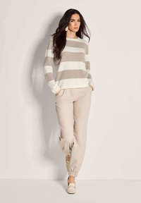Frau mit langen dunklen Haaren, die einen beigen und weißen gestreiften Pullover, beige Jogginghosen und cremefarbene Slipper trägt und vor einem hellen Hintergrund steht.