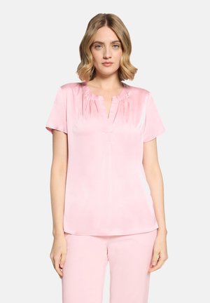 Frau mit blondem Haar, die eine hellrosa Satin-Bluse mit kurzen Ärmeln und passende rosa Hosen vor einem einfarbigen Hintergrund trägt.