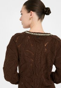 Kobieta z warkoczem w koku nosi brązowy sweter o grubej dzianinie z metaliczną dekoracyjną lamówką wzdłuż dekoltu, zwrócona w bok.