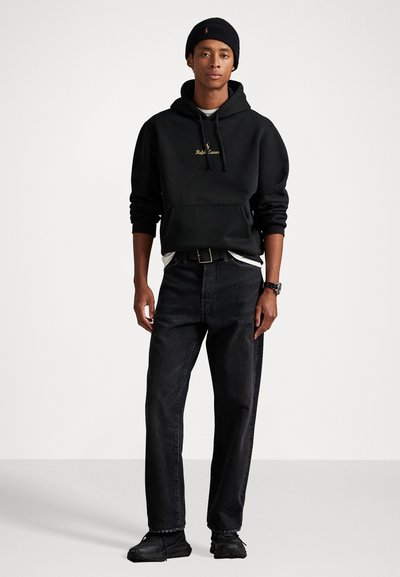 Jauns vīrietis, ģērbies melnā cepurē, melnā kapucē ar zelta Ralph Lauren logo, melnās biksēs, melnā jostā, skatīties un melnās kedās, stāv.