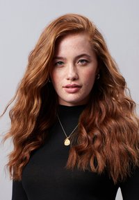 Model mit langen, welligen roten Haaren, trägt einen schwarzen Rollkragenpullover und eine goldene Anhänger-Kette, vor einem schlichten Hintergrund. Sommersprossen sind im Gesicht sichtbar.