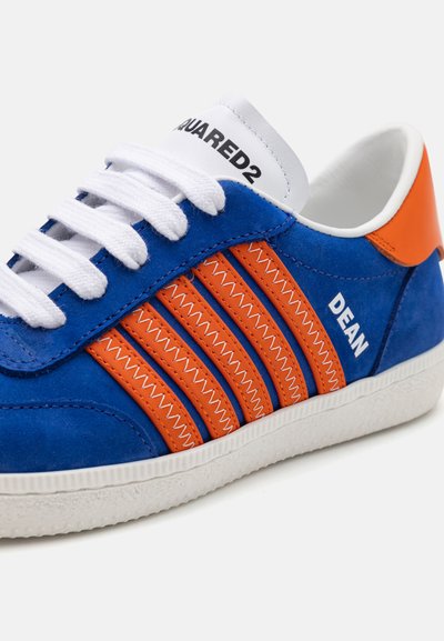 Zapatilla de ante azul con acentos naranjas y tres rayas paralelas. Presenta cordones blancos y una suela de goma texturizada. La etiqueta dice "DEAN."