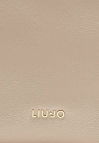 Svetlo béžová textúrovaná kožená pozadia s embosovaným zlatým logom "LIU·JO" v spodnej časti.