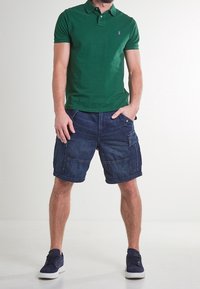 Grön polo, denim cargo-shorts och marinblå sneakers. Polon har krage och logotyp; shortsen har flera fickor.