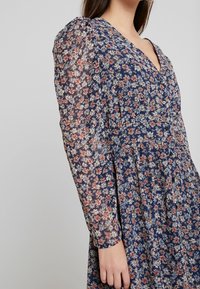 Bloemenjurk in marineblauw met lange, doorschijnende mouwen. Voorzien van een V-hals, kleine knoopjes en een geplooide taille, versierd met kleurrijke bloemen.