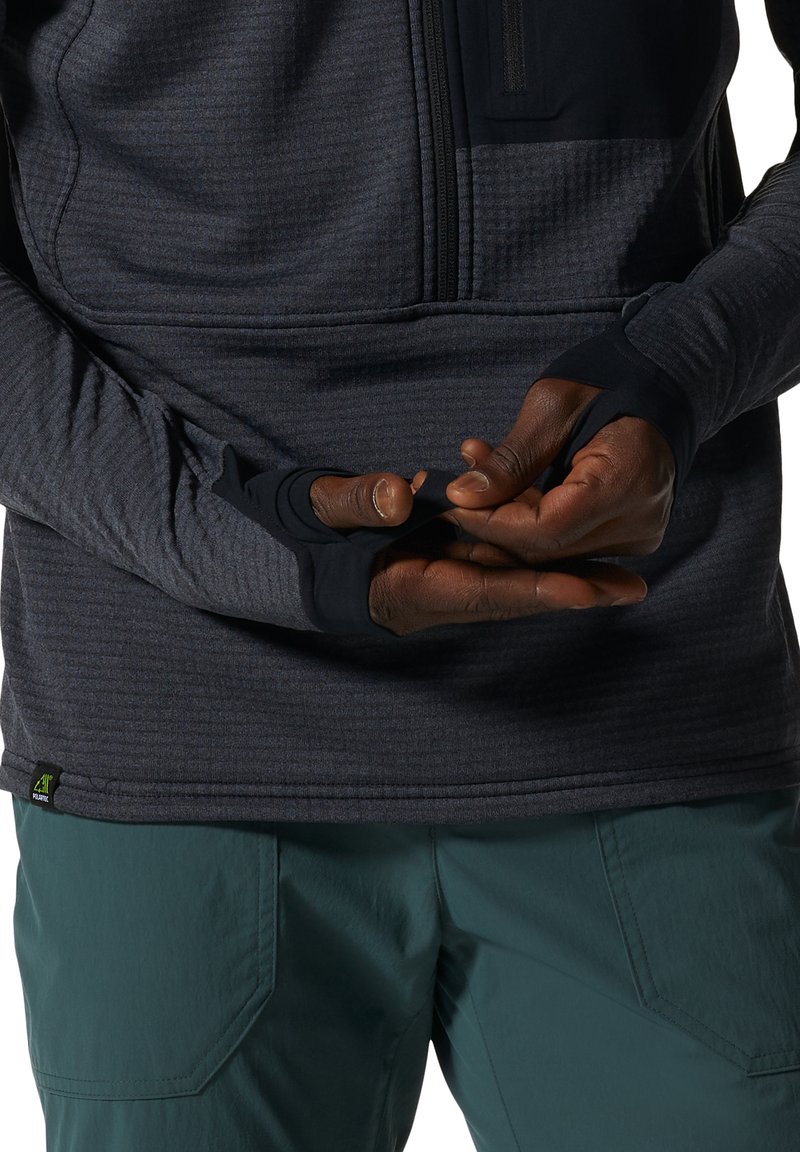 Mężczyzna noszący ciemnoszary, teksturowany, długi rękaw z otworami na kciuki naciąga materiał na niebiesko-zielone spodnie.