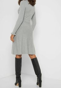 Robe grise à côtes avec des manches longues et une jupe évasée ; présente des accents de volants subtils au niveau des poignets et une taille cintrée. Bottes noires montantes.