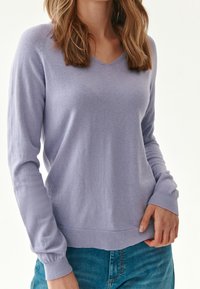 Jasnofioletowy sweter w serek z długimi rękawami i prążkowanymi mankietami. Miękka dziana tekstura, noszony na dżinsach z delikatnym detalem logo.