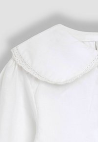 Weißes Kleidungsstück mit einem breiten, abgerundeten Kragen, der mit Spitzendetails versehen ist, aus weichem Stoff gefertigt, aus dem Schulterbereich gezeigt.
